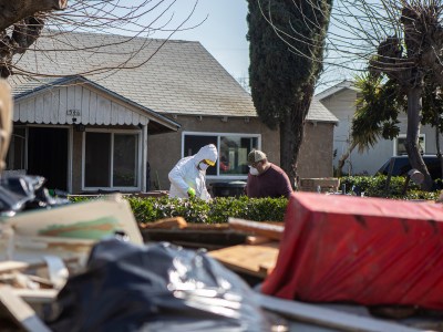 Two people in protective gear and face masks cleaning up the front year of a house with a pile of debris nearby.