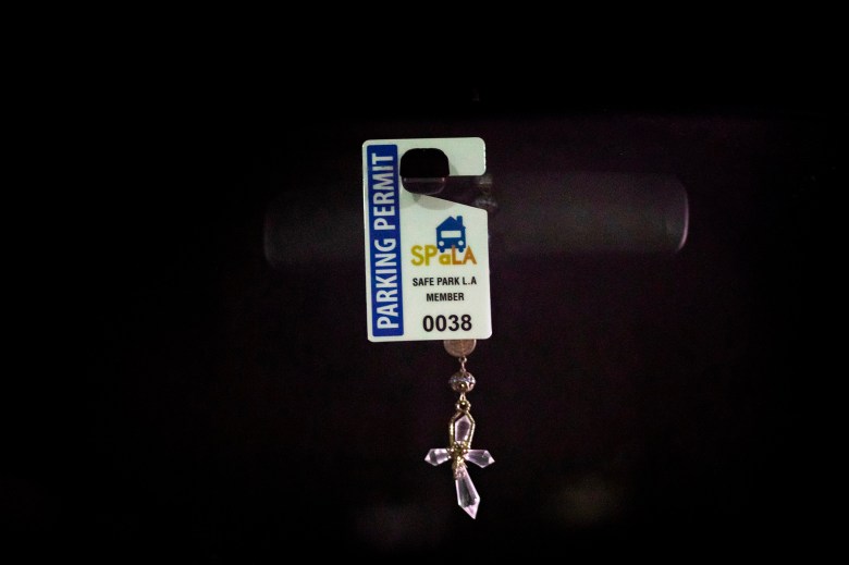 A Safe Park L.A. parking permit, labeled with the number 0038, hangs from a vehicle's rearview mirror. Attached to the permit is a delicate crystal cross, reflecting light in the dark surroundings. The permit signifies participation in the Safe Park program, which provides safe overnight parking for individuals living in their vehicles.