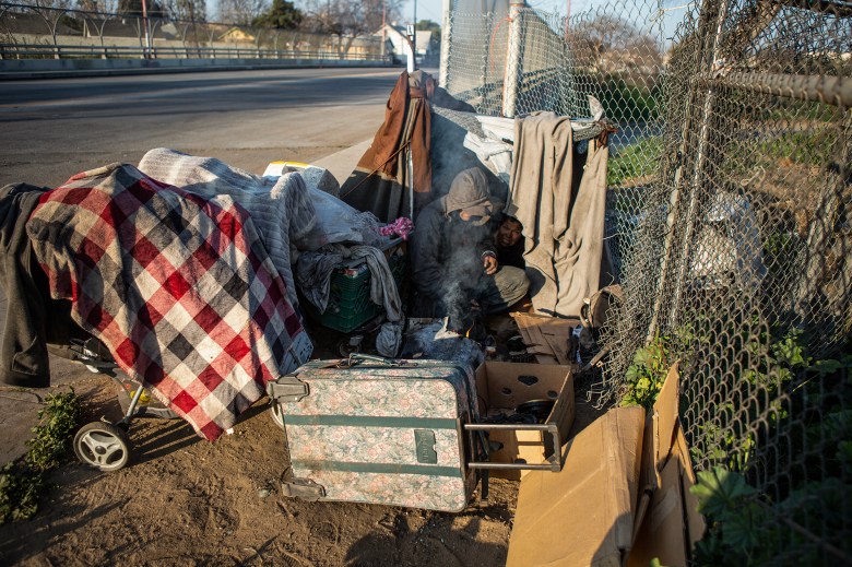 A man starts a fire while his wife sleeps inside their makeshift tent along a barbed wire fence near Highway 99 in southwest Fresno on Feb. 11, 2022. Photo by Larry Valenzuela for CalMatters/CatchLight Local