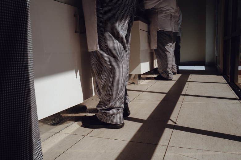 A low-angle view of individuals wearing checkered pants and aprons, standing at workstations in a well-lit space. Long shadows from a nearby window stretch across the tiled floor, suggesting late afternoon sunlight. The scene conveys a sense of quiet activity in a professional or educational setting.