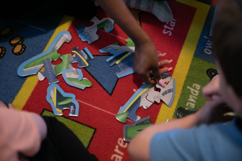 Close-up of several hands working on a colorful puzzle on a brightly patterned carpet. One hand, with neatly manicured nails, reaches for a puzzle piece, while another figure, partially visible, observes or assists. The carpet features vibrant red, blue, green, and yellow sections with printed text and designs. The atmosphere suggests collaborative play or learning.