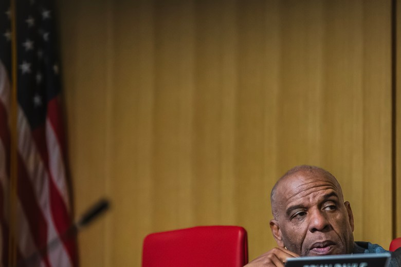 State Sen. Steven Bradford sits in a senate committee chamber. His head appears in the right-hand corner of the frame. The space above him is a wood-paneled wall. At left, an American flag rests on a vertical flagpole.