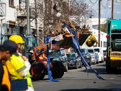 A bulldozer carrying rubble from an homeless encampment to a garbage truck parked in the street while a worker looks on.