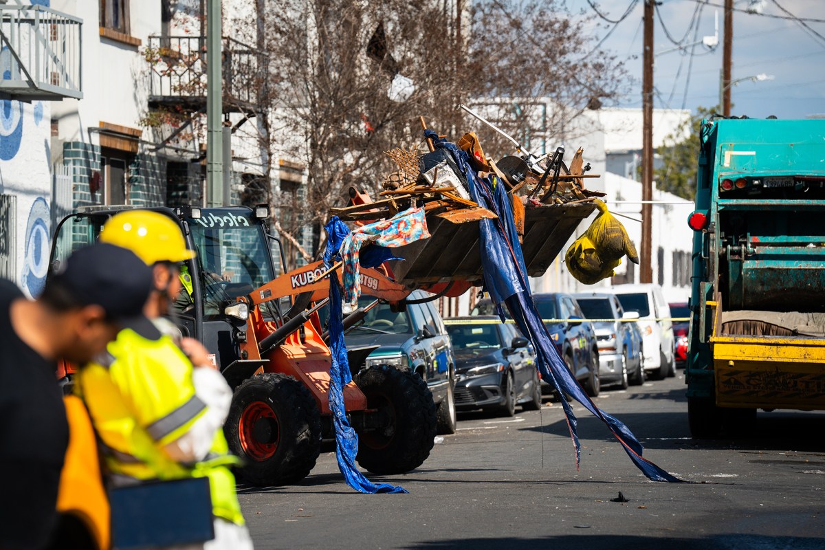 A bulldozer carrying rubble from an homeless encampment to a garbage truck parked in the street while a worker looks on.