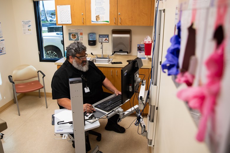 A doctor sits at a desk inside an examination room at a clinic.