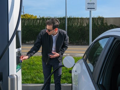 A person, wearing black sunglasses, a black sweater and blue pants, scans their credit card on an electric vehicle charging port while a pump is connected to their white car.