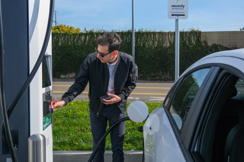 A person, wearing black sunglasses, a black sweater and blue pants, scans their credit card on an electric vehicle charging port while a pump is connected to their white car.