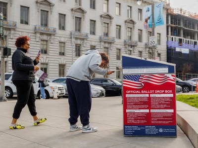 A person deposits a ballot into an official ballot drop box on a city sidewalk. Another person stands nearby, holding a phone and election pamphlet, observing the process. The ballot drop box has an American flag design with "OFFICIAL BALLOT DROP BOX" clearly visible in bold text. The backdrop shows a large, historic building with cars parked on the street.