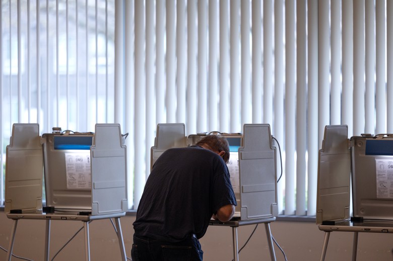 Three voting booths lined up against a wide window. One person looks down at a monitor inside the voting booth in the center.