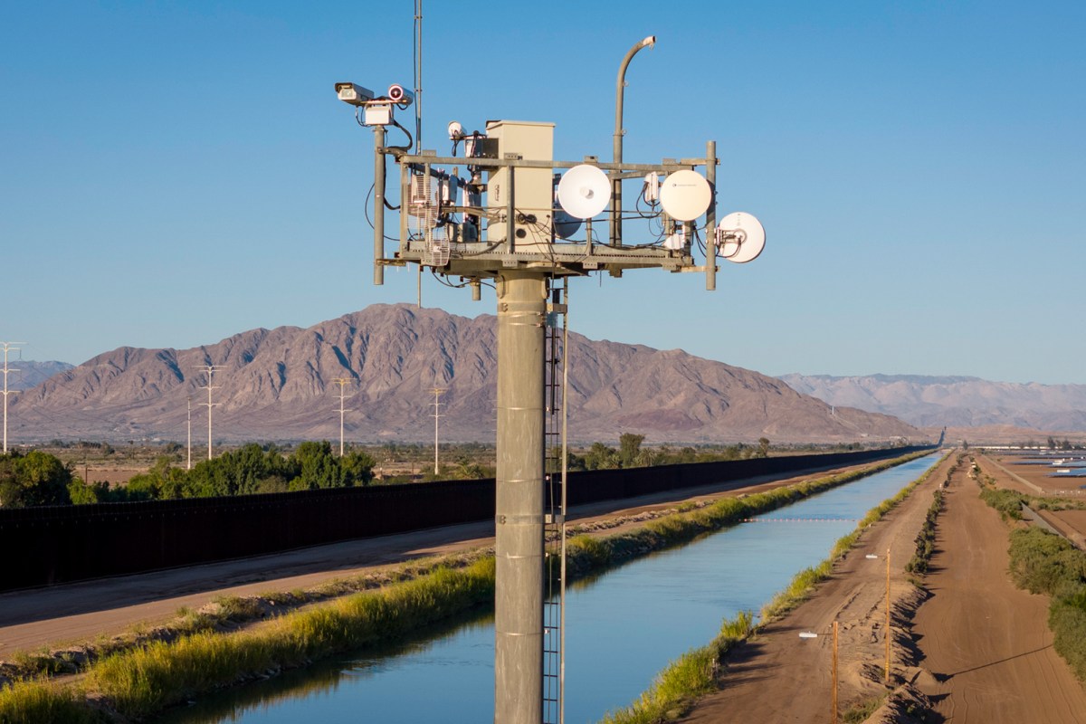 A surveillance tower monitor, with a background landscape of a brown fence, river and mountains.