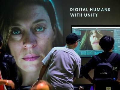 Two individuals look at a screen with an AI generated face on it, as the the words "DIGITAL HUMANS WITH UNITY" appear on the wall above the screen. To their left, another face looks out towards the space.