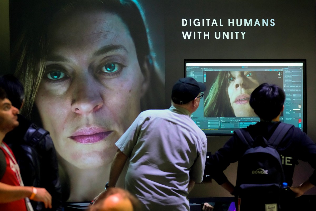 Two individuals look at a screen with an AI generated face on it, as the the words "DIGITAL HUMANS WITH UNITY" appear on the wall above the screen. To their left, another face looks out towards the space.