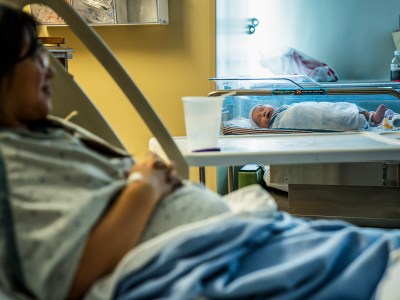 A newborn baby sleeps in a bed in a hospital room while their mother rests in a bed nearby, looking at her.