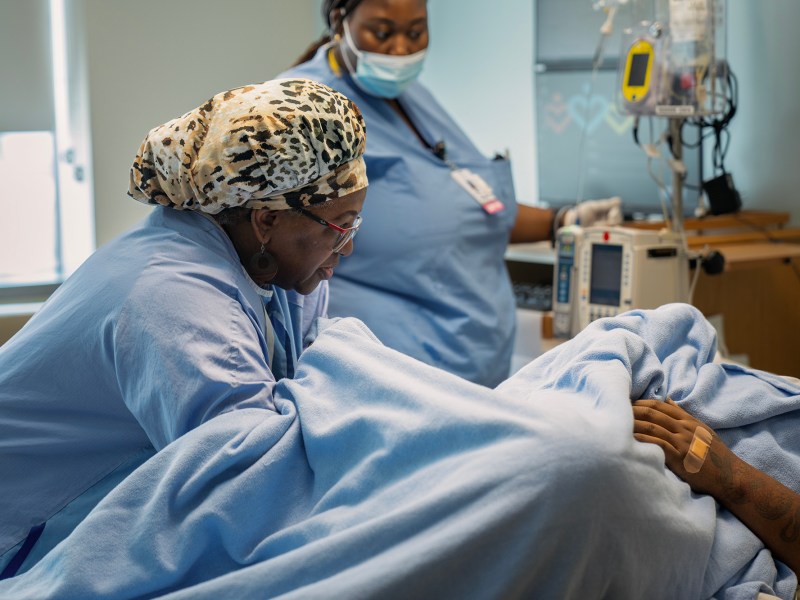 Two healthcare workers tend to a patient in a hospital bed, providing care during a medical procedure. One worker, wearing a leopard-print headscarf, closely monitors the patient while the other stands by with medical equipment. Both staff members are focused and wearing scrubs, as well as protective masks.