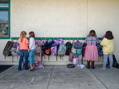 Four children stand outside a classroom against a wall with a row of hooks holding colorful backpacks. Two children on the left are facing each other, appearing to talk, while the two on the right focus on their bags. The scene captures a typical school day with bags of various colors and patterns hanging along the wall.