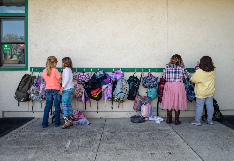 Four children stand outside a classroom against a wall with a row of hooks holding colorful backpacks. Two children on the left are facing each other, appearing to talk, while the two on the right focus on their bags. The scene captures a typical school day with bags of various colors and patterns hanging along the wall.