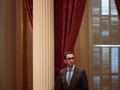 A lawmaker, wearing a tan suit and blue tie, standing in front of a podium with white pillars and red curtains in the background of the Senate Floor at the state Capitol in Sacramento.