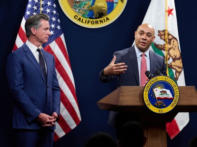 Gov. Gavin Newsom, standing to the left of the framel, listens to the director of the Department of Finance, on the right side of the frame, speak as both of them stand near a podium with an American flag, the California flag and the California state seal behind them during a press conference.