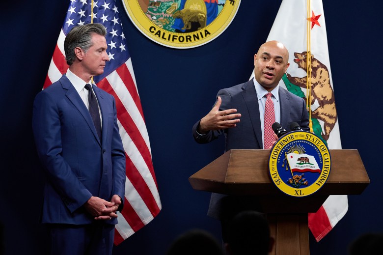 Gov. Gavin Newsom, standing to the left of the framel, listens to the director of the Department of Finance, on the right side of the frame, speak as both of them stand near a podium with an American flag, the California flag and the California state seal behind them during a press conference.