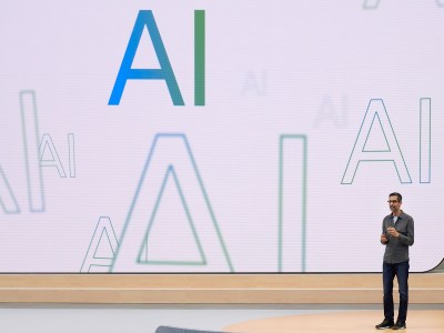 A person in a button up shirt and blue jeans stands in front of a large screen displaying colorful variations of the term AI.