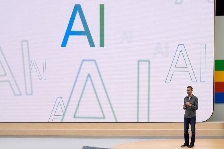 A person in a button up shirt and blue jeans stands in front of a large screen displaying colorful variations of the term AI.