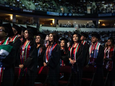 People wearing black cap and gowns with red, green and white colored sashes stand in front of chairs during a graduation ceremony in font of attendees in an arena.