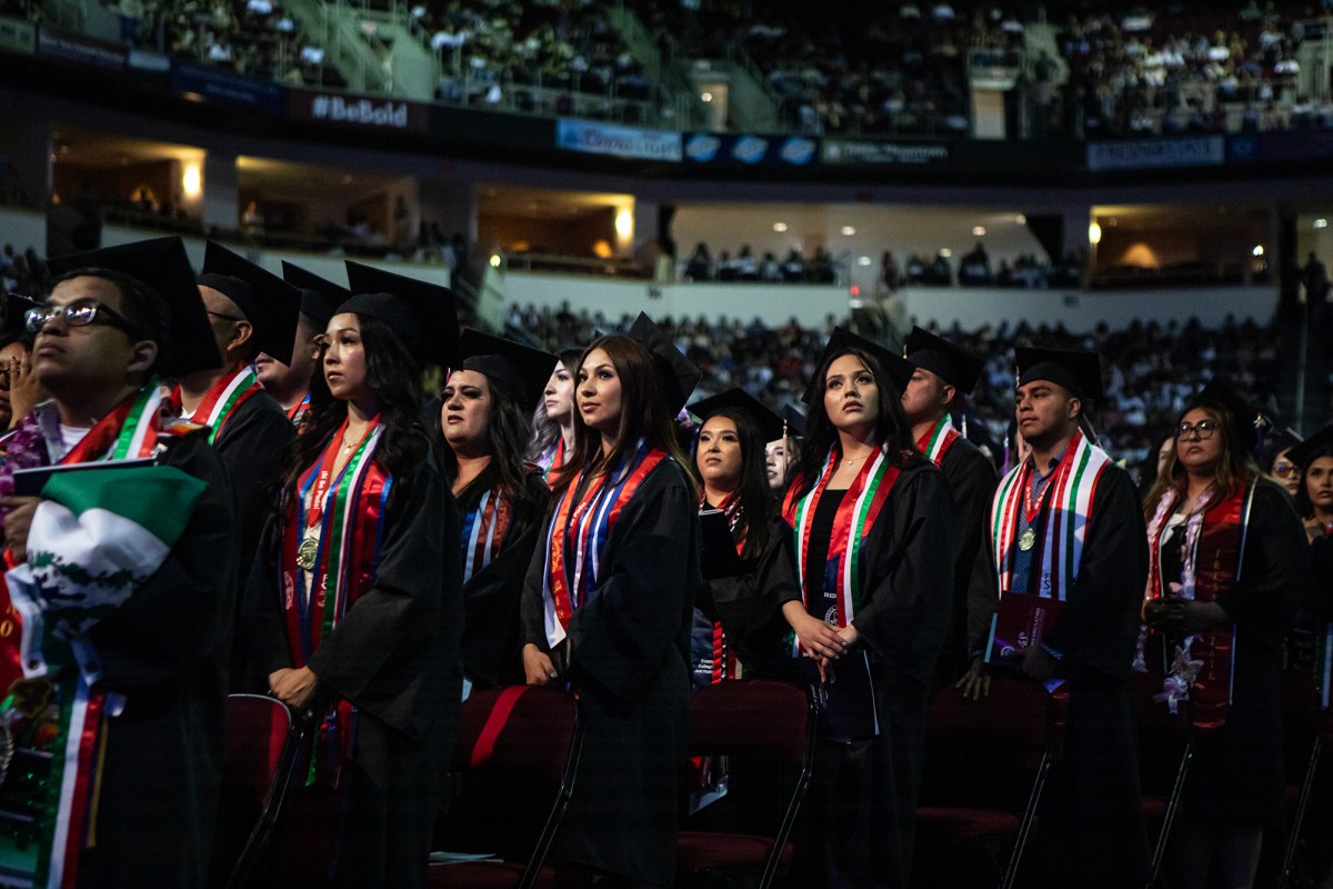 People wearing black cap and gowns with red, green and white colored sashes stand in front of chairs during a graduation ceremony in font of attendees in an arena.