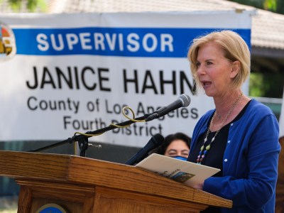 A politician, wearing a blue cardigan and black shirt, stands behind a podium with a microphone during an event at a park.