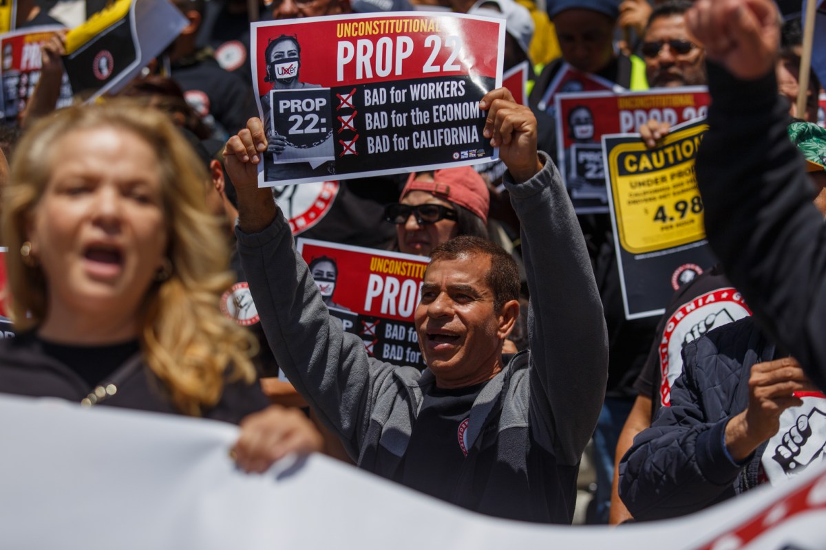 A crowd of protesters hold Prop. 22 signs with their mouths open and fists in the air. One prominent sign reads, "Unconstitutional Prop 22, Bad for Workers, Bad for the Economy, Bad for California."