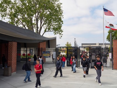 A wide view of students walking through the black gates of a school campus with brick buildings. An American and California state flag can be seen in the distance.