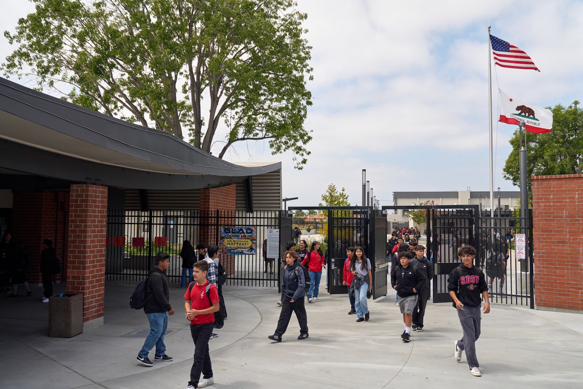 A wide view of students walking through the black gates of a school campus with brick buildings. An American and California state flag can be seen in the distance.