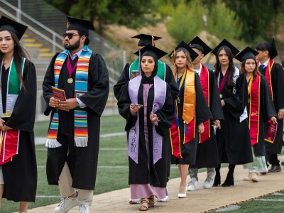 A student dressed in a graduation cap and gown with a purple satchel and tassel walks alongside other graduates at a commencement ceremony.