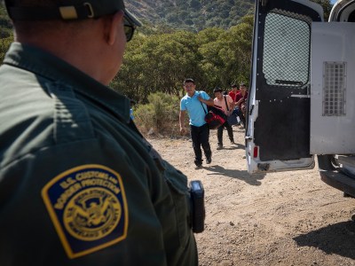 A U.S. Customs and Border Protection officer, identifiable by a patch on their uniform, stands near an open van door, observing a group of people approaching. The scene is outdoors in a rural area with dry, rugged terrain and sparse vegetation. The individuals walking towards the van carry personal belongings and appear to be guided or escorted by the officer.