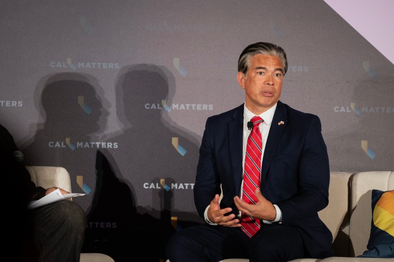 A person in a dark blue suit and a red tie sits in a chair on stage and gestures while they speak. At left, the person's shadow, and that of a moderator are visible on a backdrop with the words "CALMATTERS" emblazoned on it. The setting is a forum or panel.