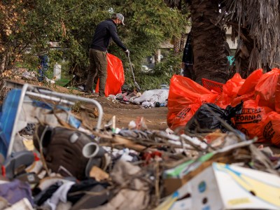 Gov. Gavin Newsom, dressed is brown pants, a black jacket and a grey baseball hat, picks up trash from a homeless encampment.