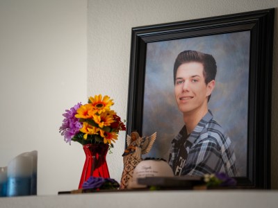 A framed portrait of a smiling person sits on a shelf. Next to the frame is a vibrant bouquet of yellow, orange, and purple flowers in a red vase, along with a small angel figurine and candles. The setting appears to be a memorial display.