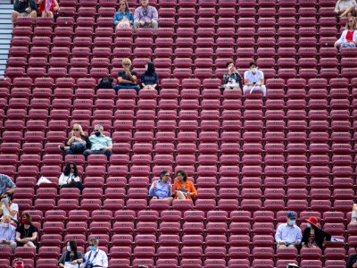 Maroon bleachers fill the frame with people, mostly in pairs, sitting throughout the bleachers with a large amount of distance between them.