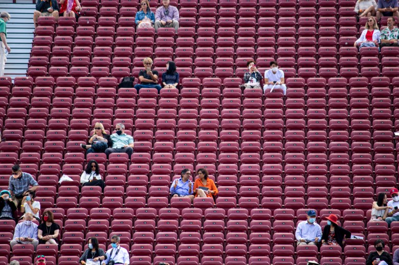 Maroon bleachers fill the frame with people, mostly in pairs, sitting throughout the bleachers with a large amount of distance between them.