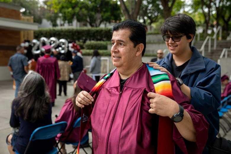 Marcelo Baca, 62, puts on a graduation robe with the help of his son, Jason, 16, before a graduation ceremony at Mt. San Antonio College in Walnut on June 15, 2023. Photo by Alisha Jucevic for CalMatters