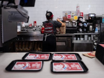 A person in a kitchen at a fast-food restaurant prepares a meal, while in the foreground, four trays sit on a counter.