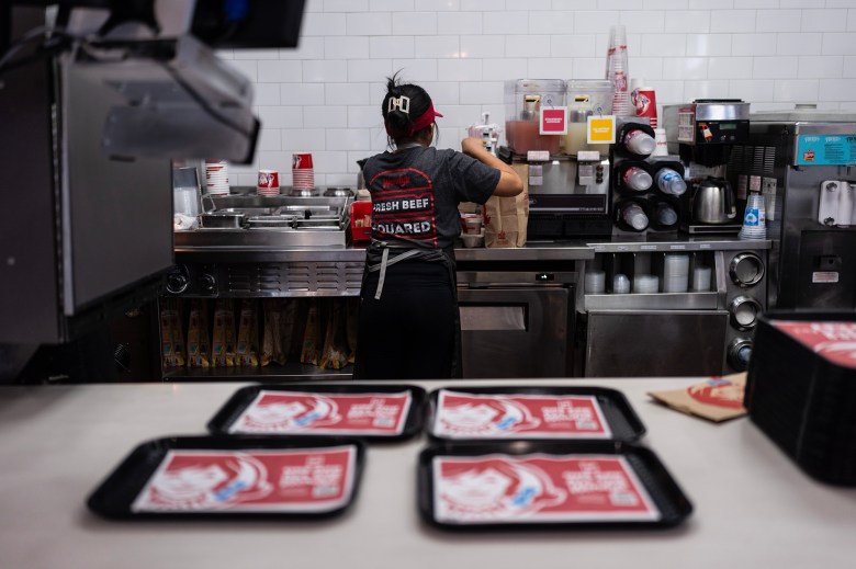 A person in a kitchen at a fast-food restaurant prepares a meal, while in the foreground, four trays sit on a counter.