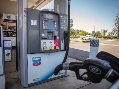 A gas pump at a Chevron station displays a screen showing $185.71 spent and 29.482 gallons pumped. The pump offers three fuel options: Regular (87), Plus (89), and Supreme (91). A gas nozzle is visible in the foreground, resting in its holder. In the background, a road with a truck passing by and trees can be seen. A nearby pump station includes an advertisement for a mobile app and fuel rewards.