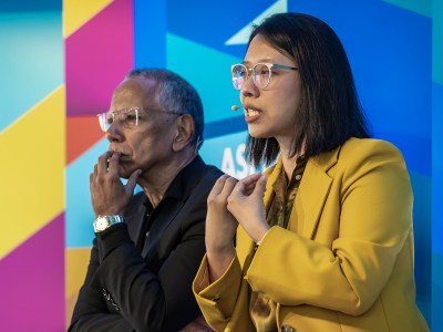 Two people are sitting side by side against a colorful geometric background. The person on the right is speaking, wearing a yellow blazer and clear-framed glasses, and gesturing with their hands. They have a microphone attached to their face. The person on the left, wearing a black jacket, is resting their chin on one hand, appearing thoughtful. Both are engaged in a discussion