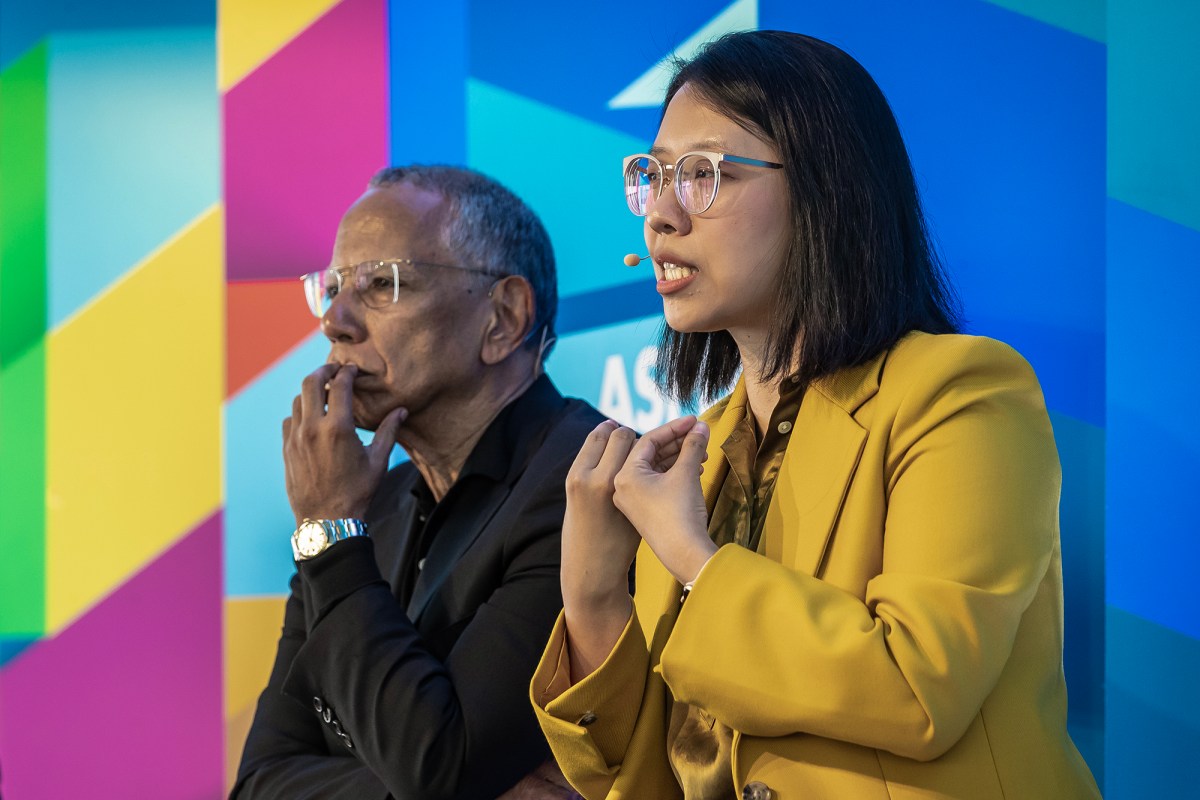 Two people are sitting side by side against a colorful geometric background. The person on the right is speaking, wearing a yellow blazer and clear-framed glasses, and gesturing with their hands. They have a microphone attached to their face. The person on the left, wearing a black jacket, is resting their chin on one hand, appearing thoughtful. Both are engaged in a discussion