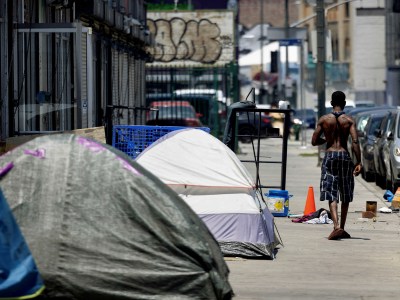 A view of a person's back as they walk by gray and white tents on the sidewalk on a hot day in Los Angeles.