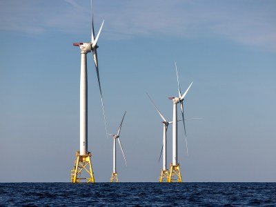 A group of offshore wind turbines rises from the ocean, with tall white towers and large three-blade rotors spinning slowly against a clear blue sky. The turbines are anchored on yellow platforms, standing above the water. The horizon stretches across the image, showcasing the expansive open sea and a sense of renewable energy in action.