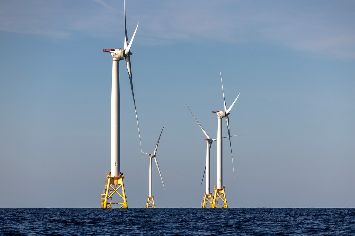 A group of offshore wind turbines rises from the ocean, with tall white towers and large three-blade rotors spinning slowly against a clear blue sky. The turbines are anchored on yellow platforms, standing above the water. The horizon stretches across the image, showcasing the expansive open sea and a sense of renewable energy in action.