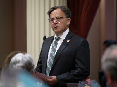 A person dressed in a black suit, white button-up shirt and a black and green tie speaks into a microphone while holding a piece of paper. The heads of other people are visible at the bottom of the frame, as they listen. The setting is a legislative hearing.