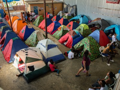 A large indoor shelter filled with colorful tents, where people have set up temporary living spaces on the concrete floor. Some individuals are sitting outside their tents or on chairs along the wall, while others move around the area. The tents vary in colors and designs, with some families gathered together, indicating a communal space for shelter and respite.