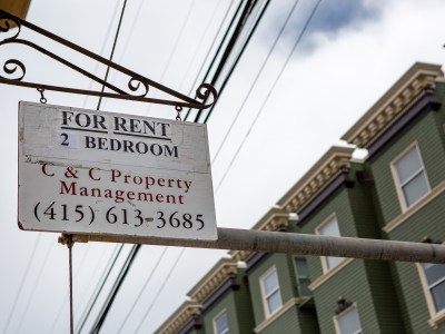 A for rent sign for a two bedroom apartment hanging off a wall of an apartment complex.